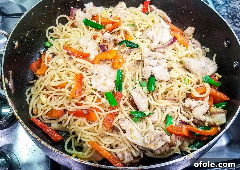 Chicken, bell peppers, and scrambled eggs being stir-fried with cooked spaghetti noodles in a large pan.
