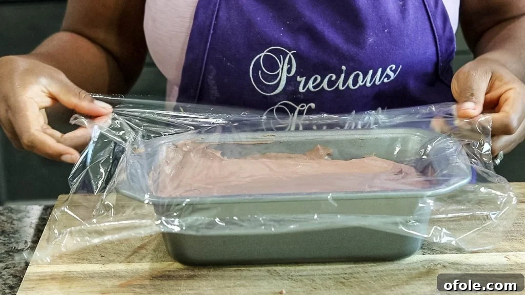 A loaf pan filled with chocolate ice cream mixture, carefully covered with plastic wrap pressed directly onto the surface to prevent ice crystals.