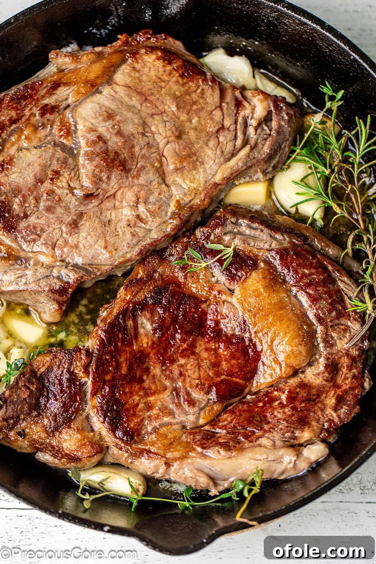 Pan Seared Ribeye Steaks in Cast Iron Two sizzling ribeye steaks in a cast iron skillet, garnished with fresh rosemary, thyme, and garlic cloves.