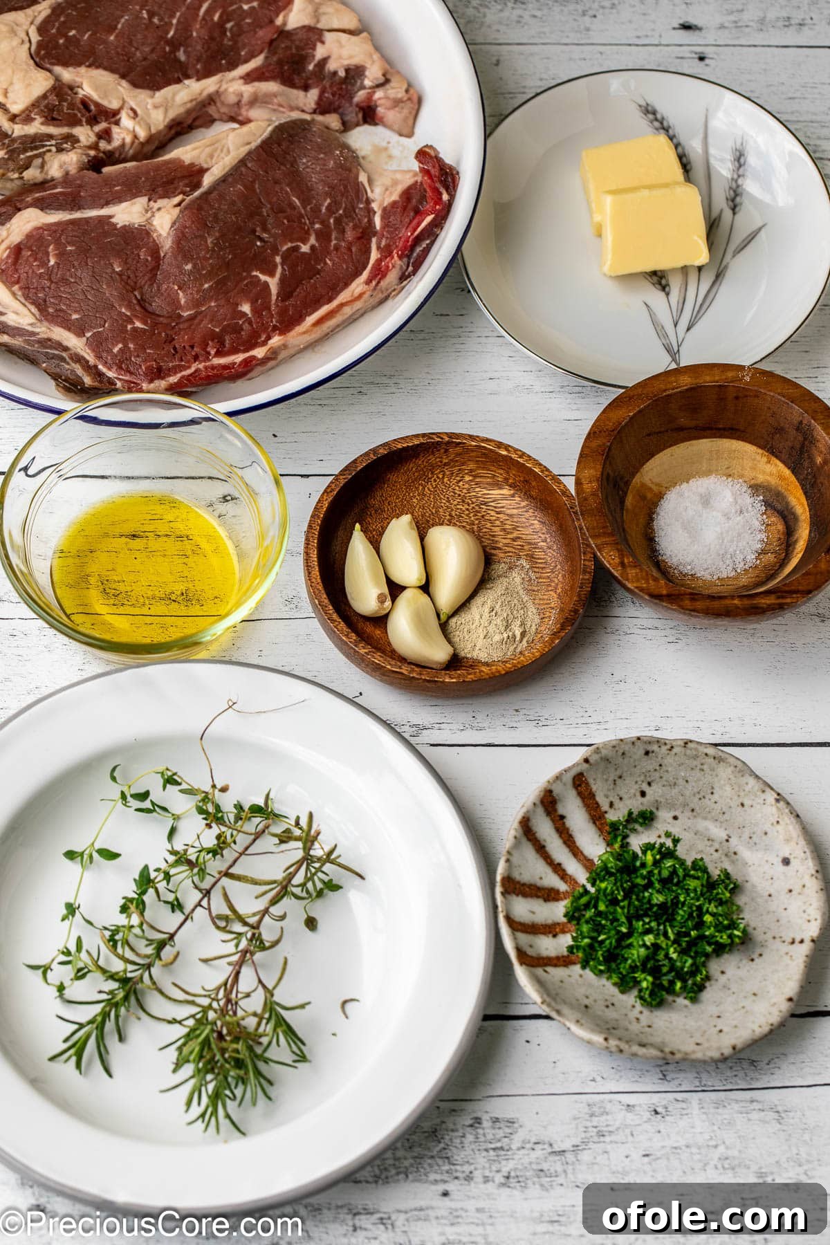 Fresh Ingredients for Ribeye A collection of fresh ingredients laid out for pan-searing steak, including ribeye, olive oil, salt, pepper, garlic, rosemary, thyme, and butter.