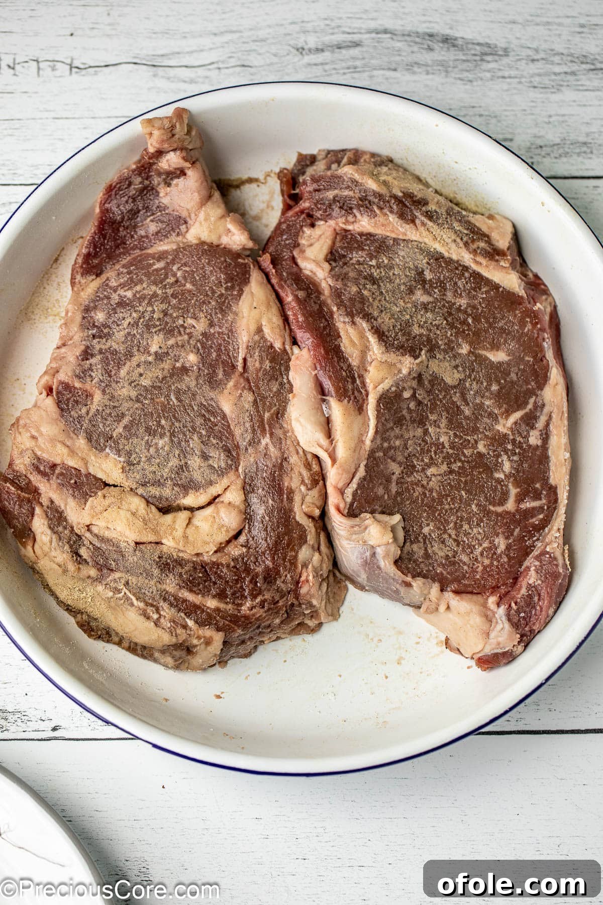Seasoned Ribeye Steaks Two seasoned ribeye steaks resting in a white bowl, ready for pan-searing.