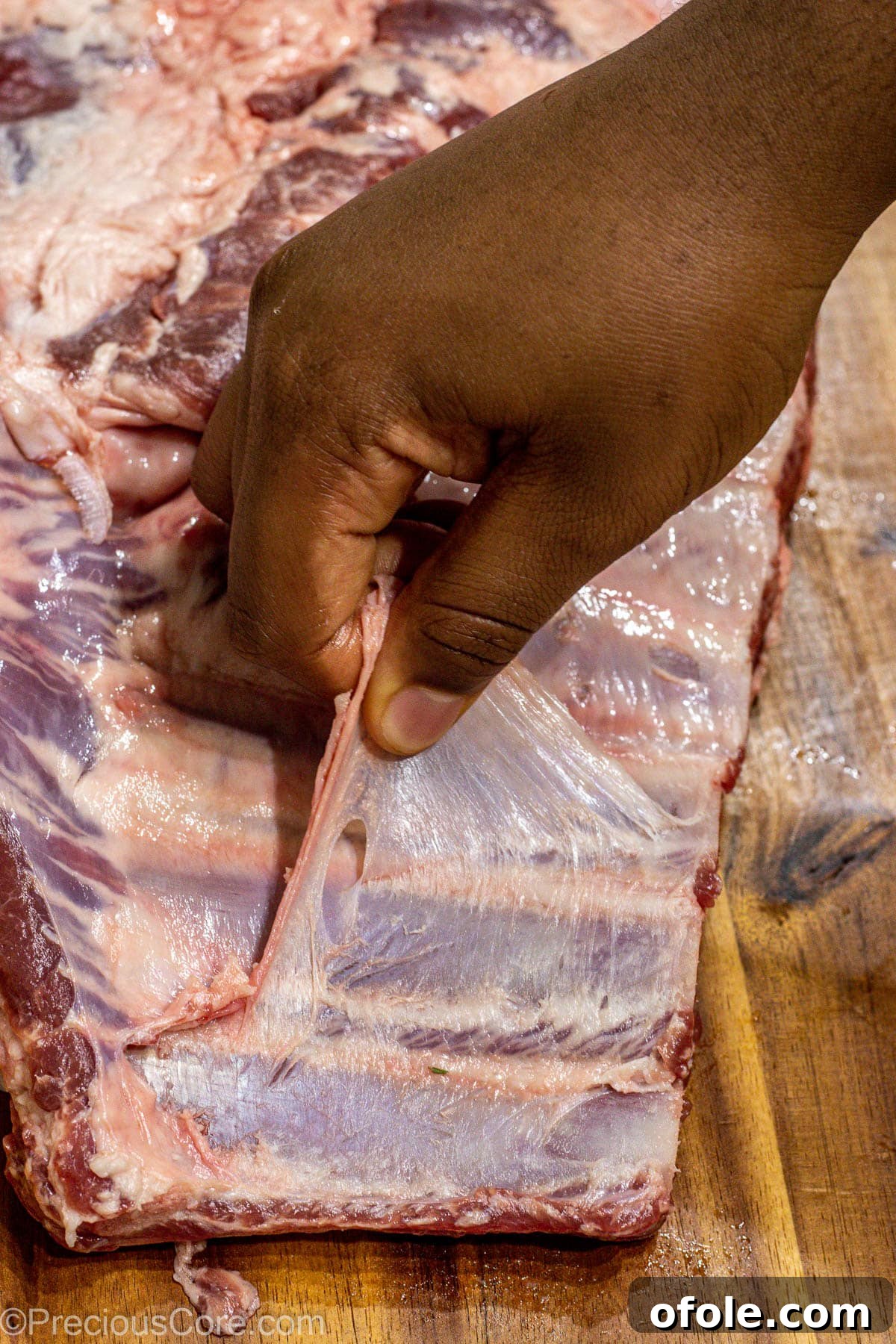 A hand expertly peeling off the thin membrane from the bone-side of a rack of spare ribs on a cutting board, preparing them for seasoning.