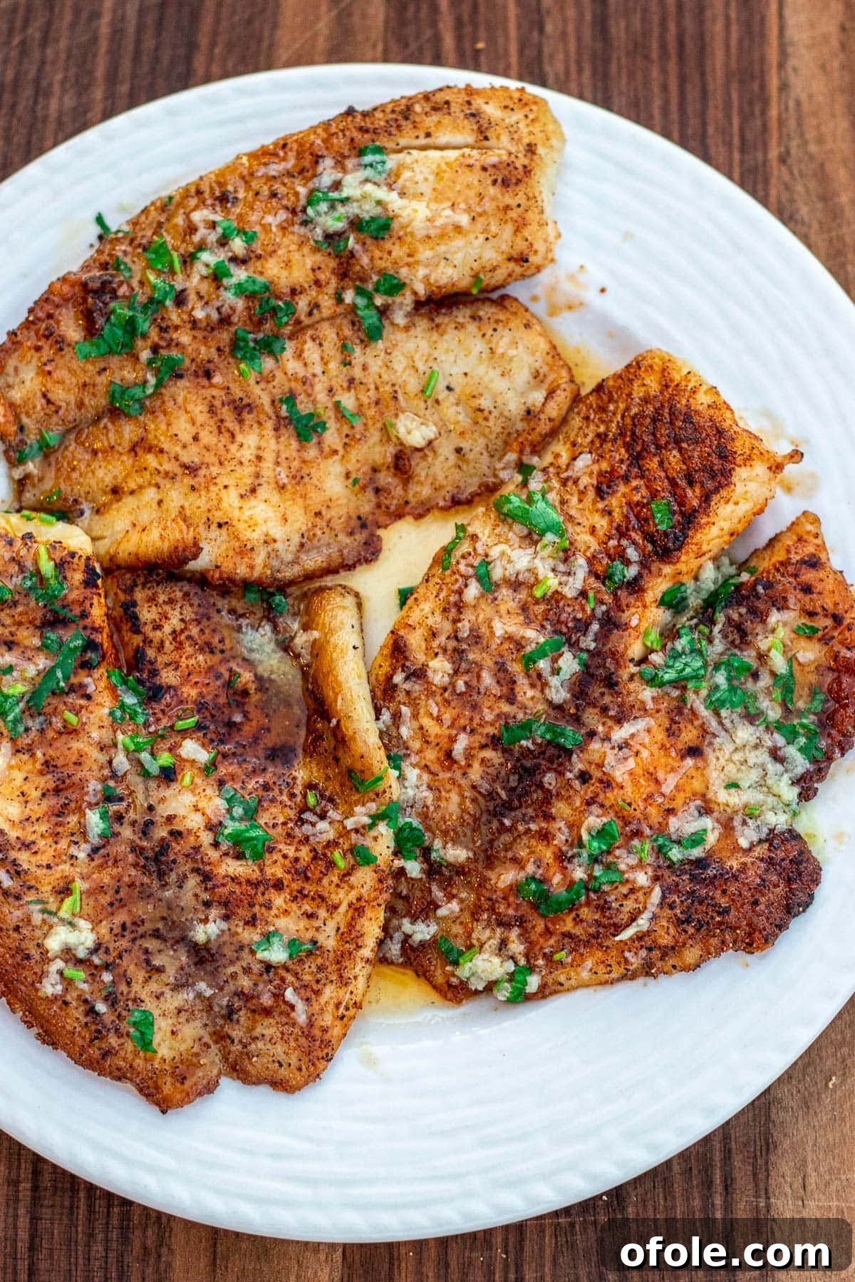 Lemon butter tilapia fillets on a white plate, garnished with fresh parsley and lemon wedges.