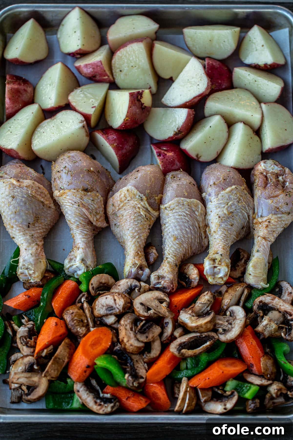 One-Pan Roasted Chicken, Potatoes, and Vegetables 5 A baking sheet is shown lined with parchment paper, holding raw, seasoned chicken drumsticks, potato chunks, and mixed vegetables, all ready for the oven.
