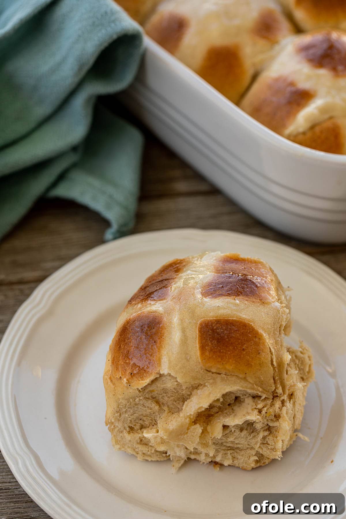 The Ultimate Hot Cross Buns 3 A single sweet spiced bun, perfectly round with a distinct white cross, resting on a rustic plate.