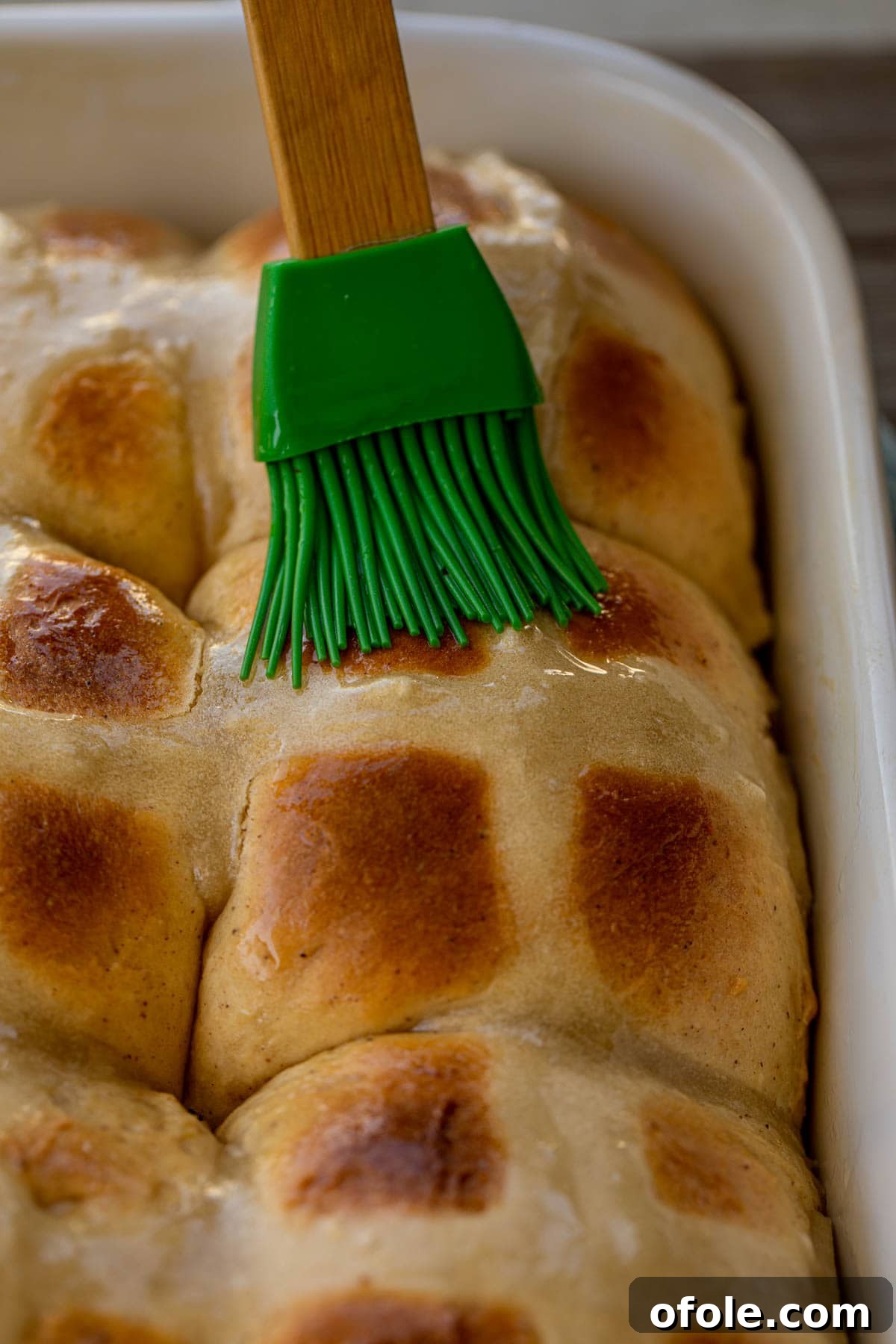 The Ultimate Hot Cross Buns 5 A pastry brush applying a shimmering glaze over freshly baked hot cross buns in a baking dish.