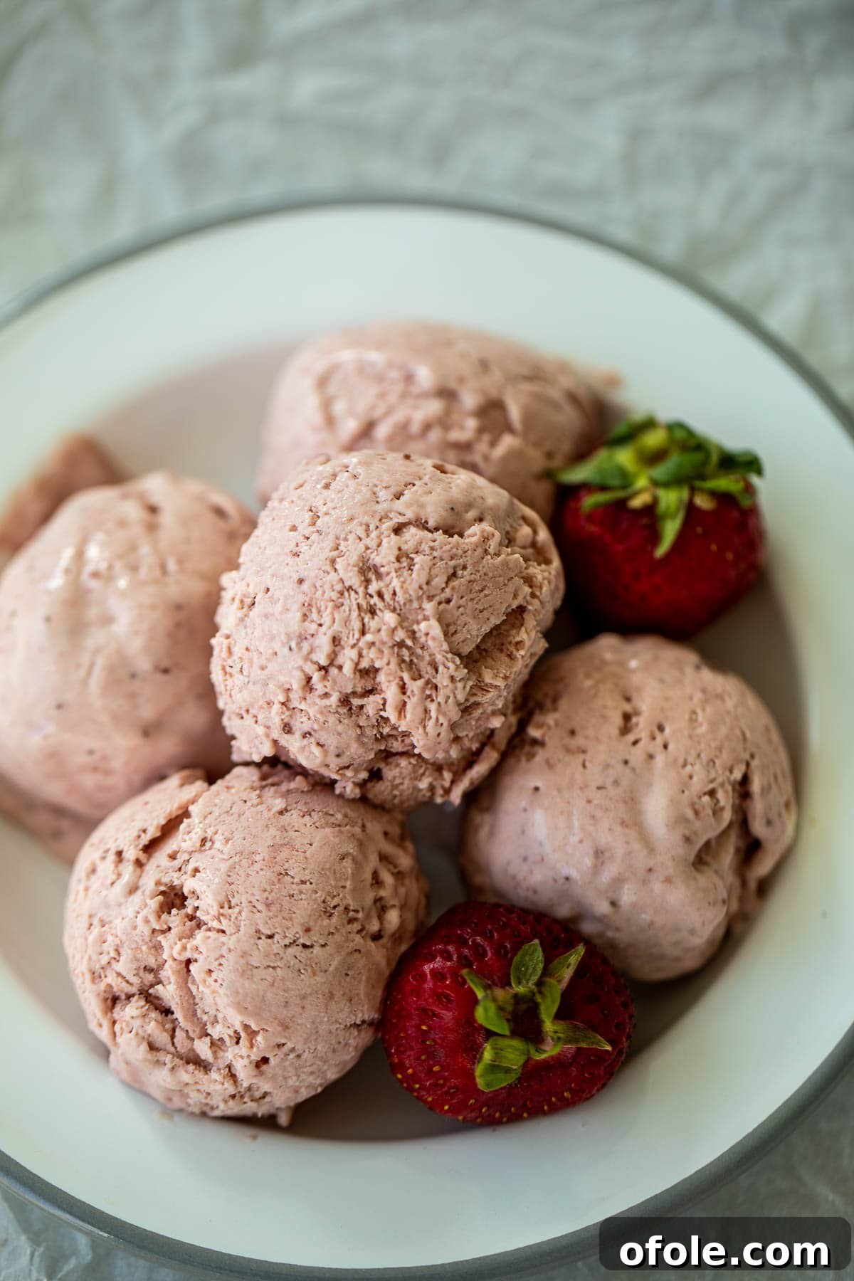 Multiple scoops of strawberry ice cream on a white plate, accompanied by a fresh strawberry and mint leaves.