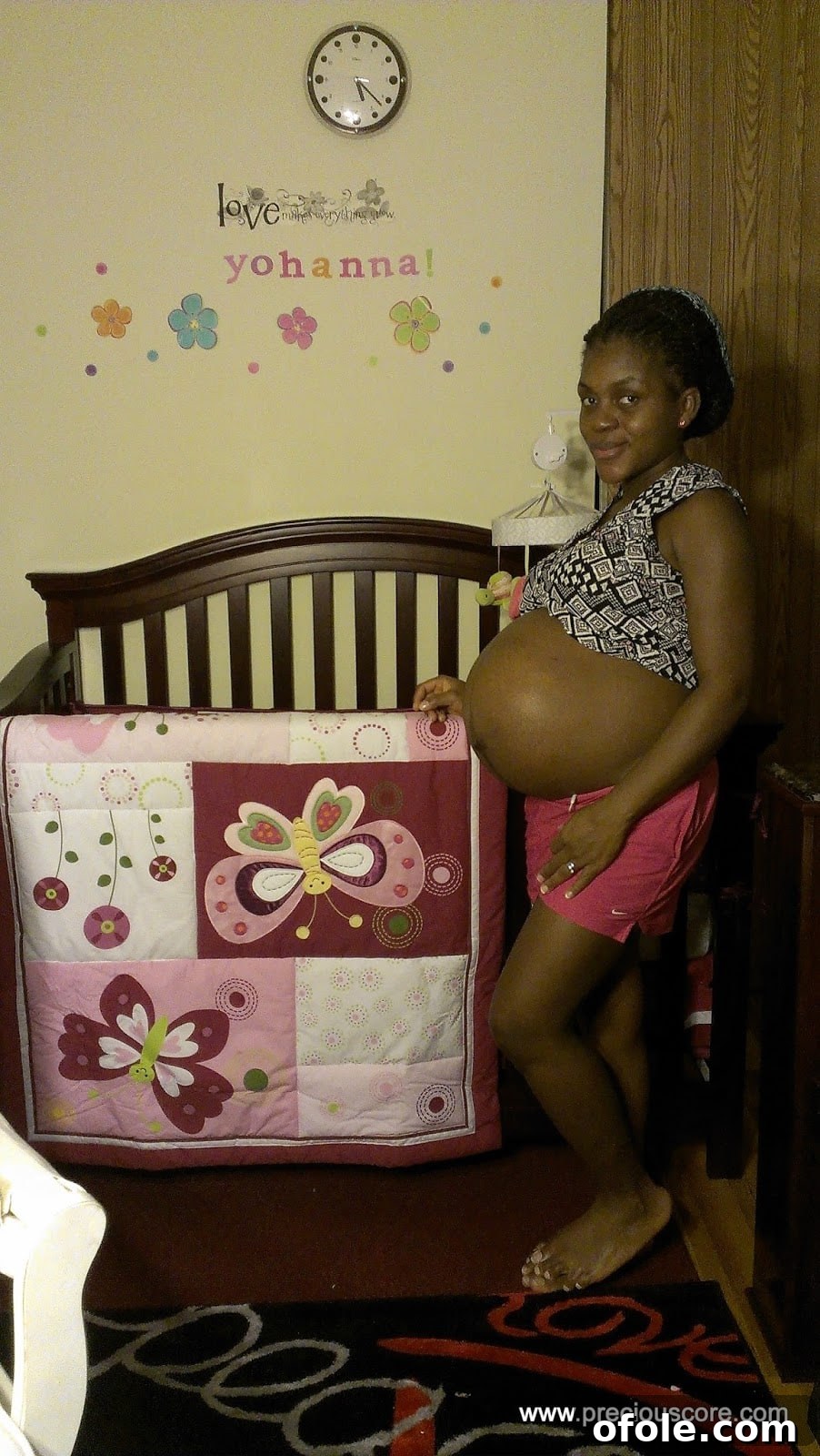 A pregnant woman standing next to a baby crib, her large belly prominent.