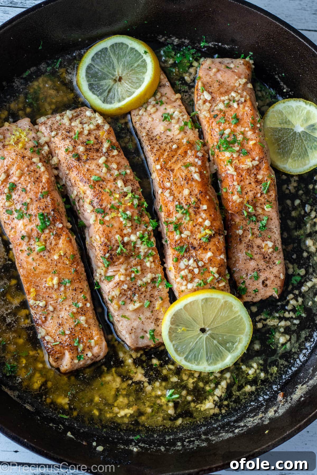 Four perfectly seared lemon butter salmon fillets in a cast iron skillet, garnished with fresh herbs.