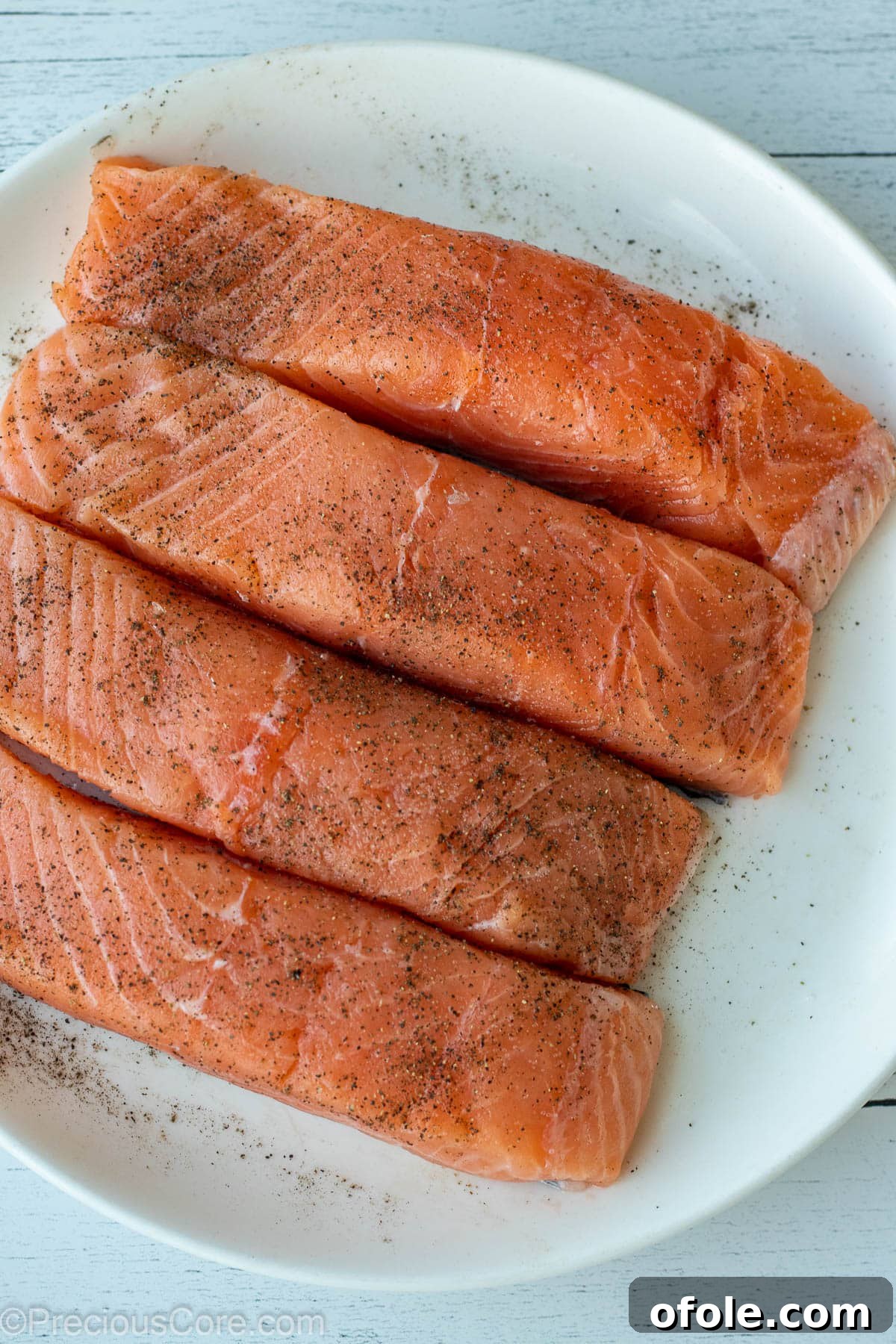 Four raw salmon fillets on a white plate, seasoned with salt and pepper, ready for searing.