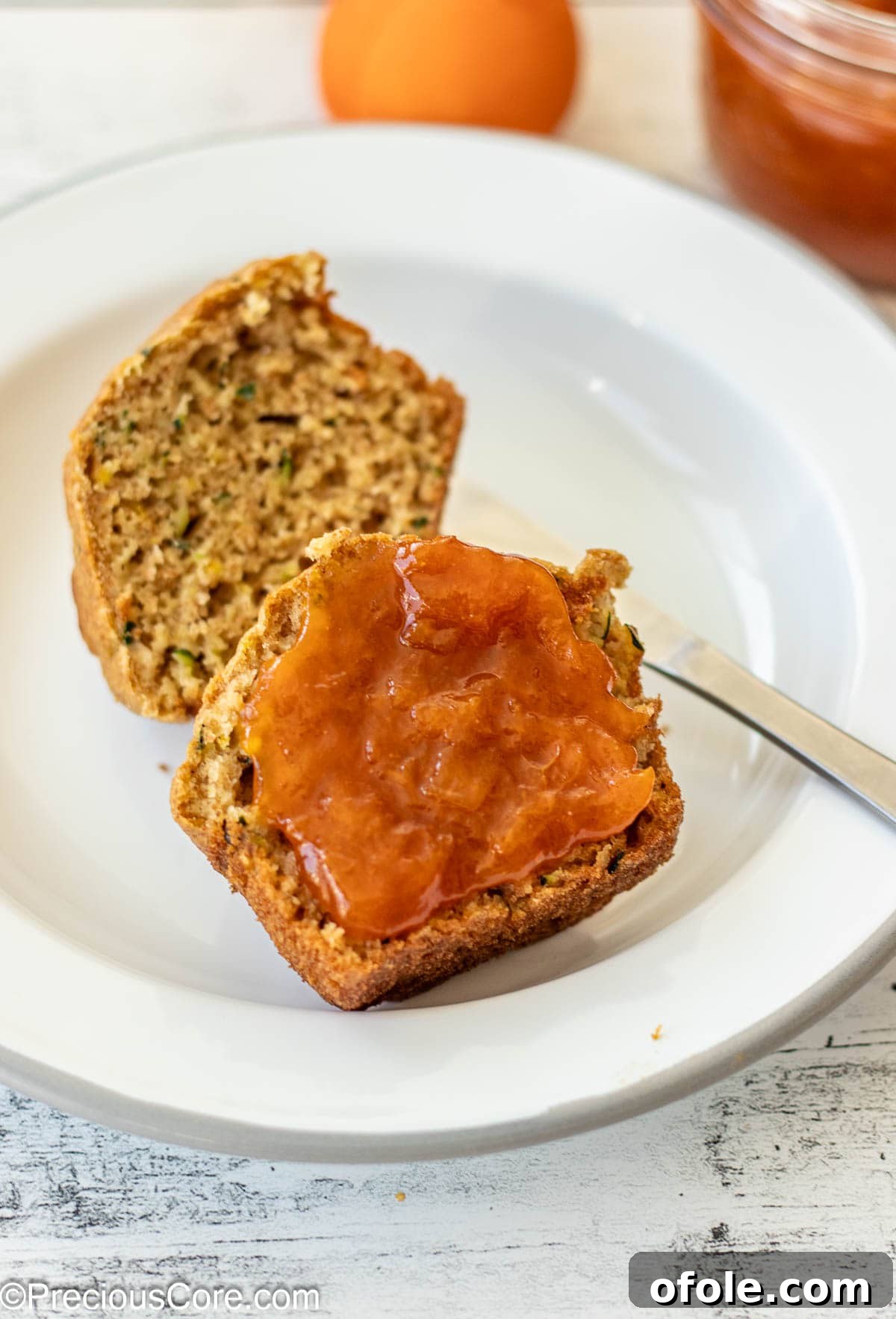 A zucchini muffin, sliced in half, with a generous spread of homemade apricot preserves on one side, showcasing its delicious texture.