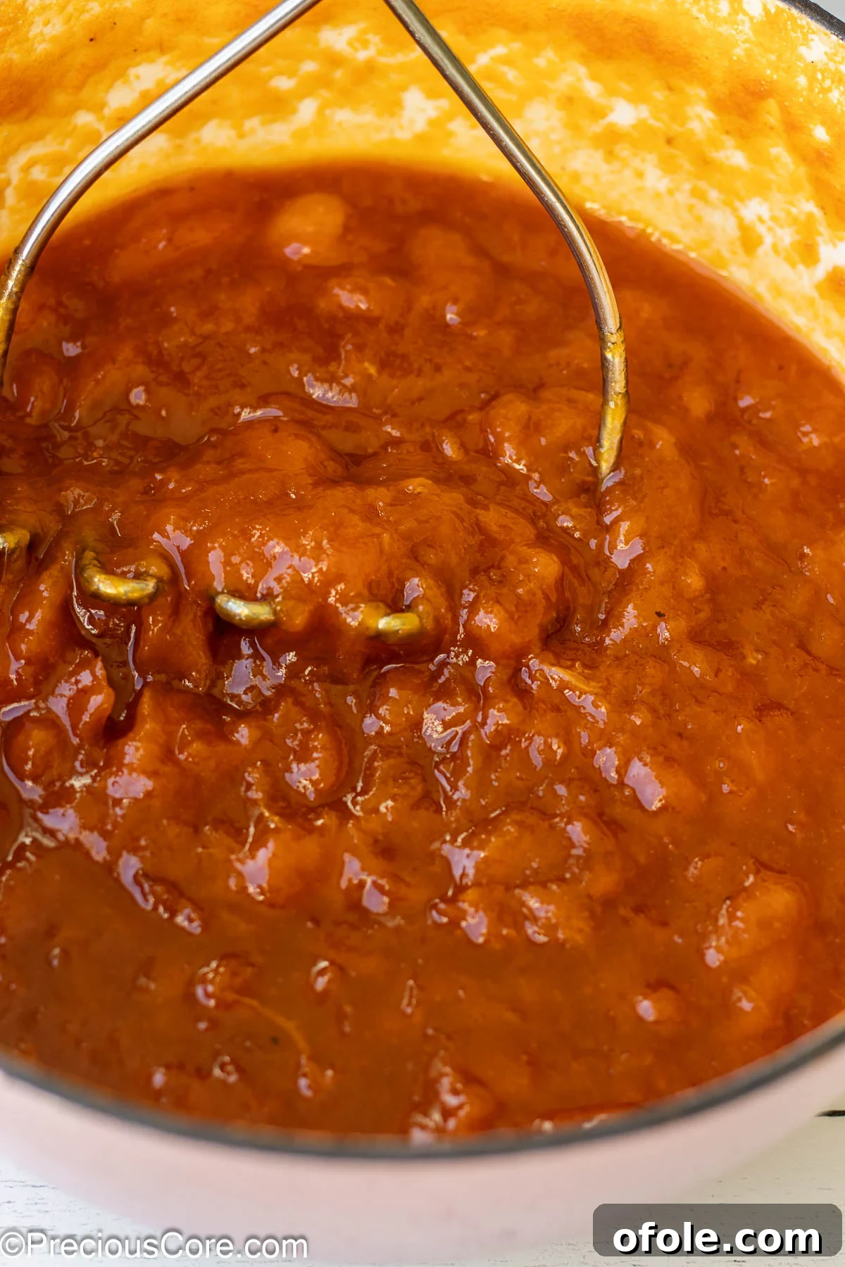 A hand using a potato masher to gently mash apricot preserves in a pot, demonstrating how to achieve a smoother consistency.