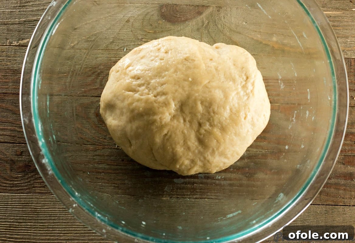 Sweet dinner roll dough in a clear glass bowl after initial mixing, showing its soft consistency.