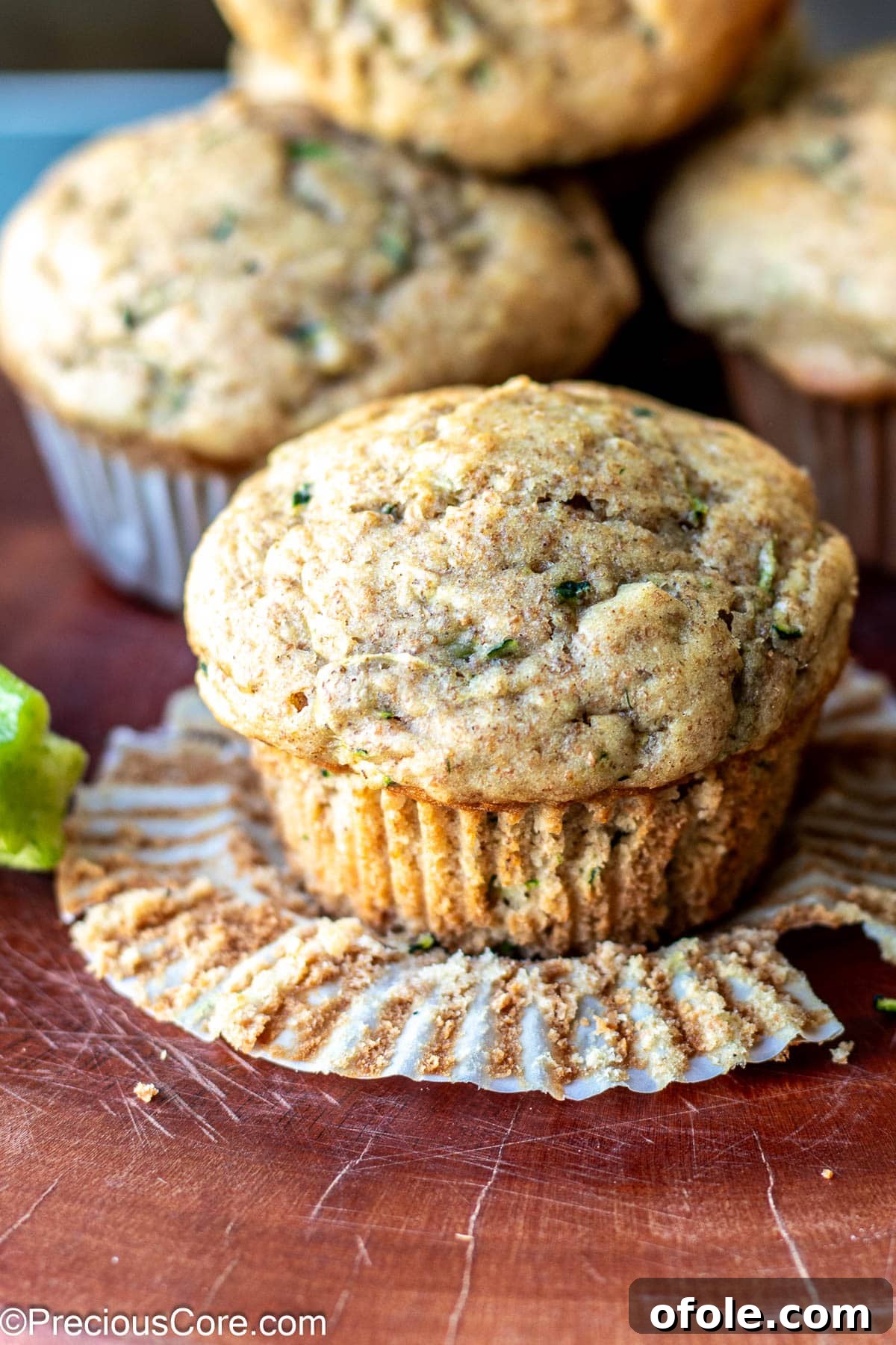 A freshly baked zucchini muffin in the foreground, with its paper lining partly peeled, revealing its golden-brown crust.