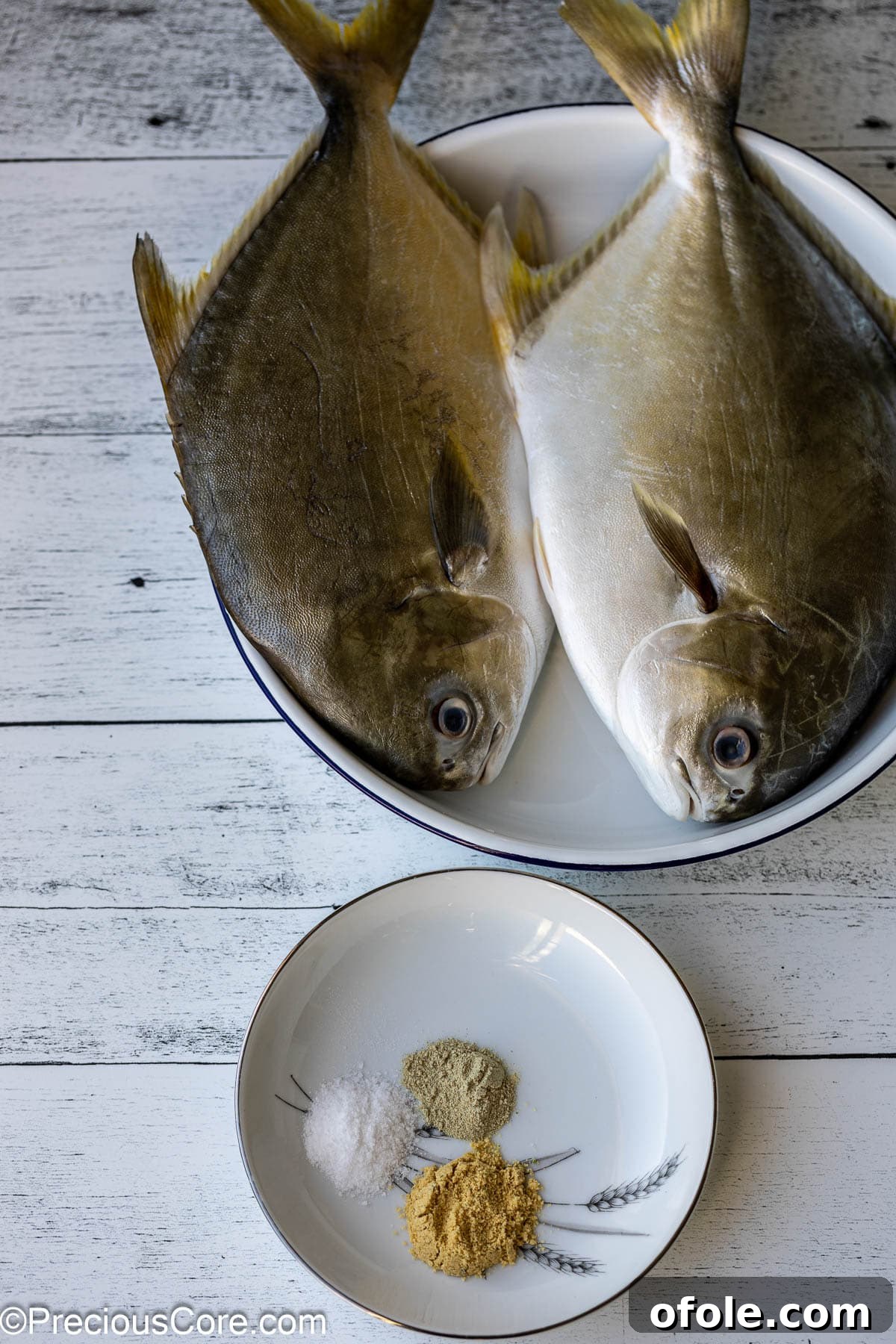 Two whole fish in a white bowl with a small plate of seasoning beside them.