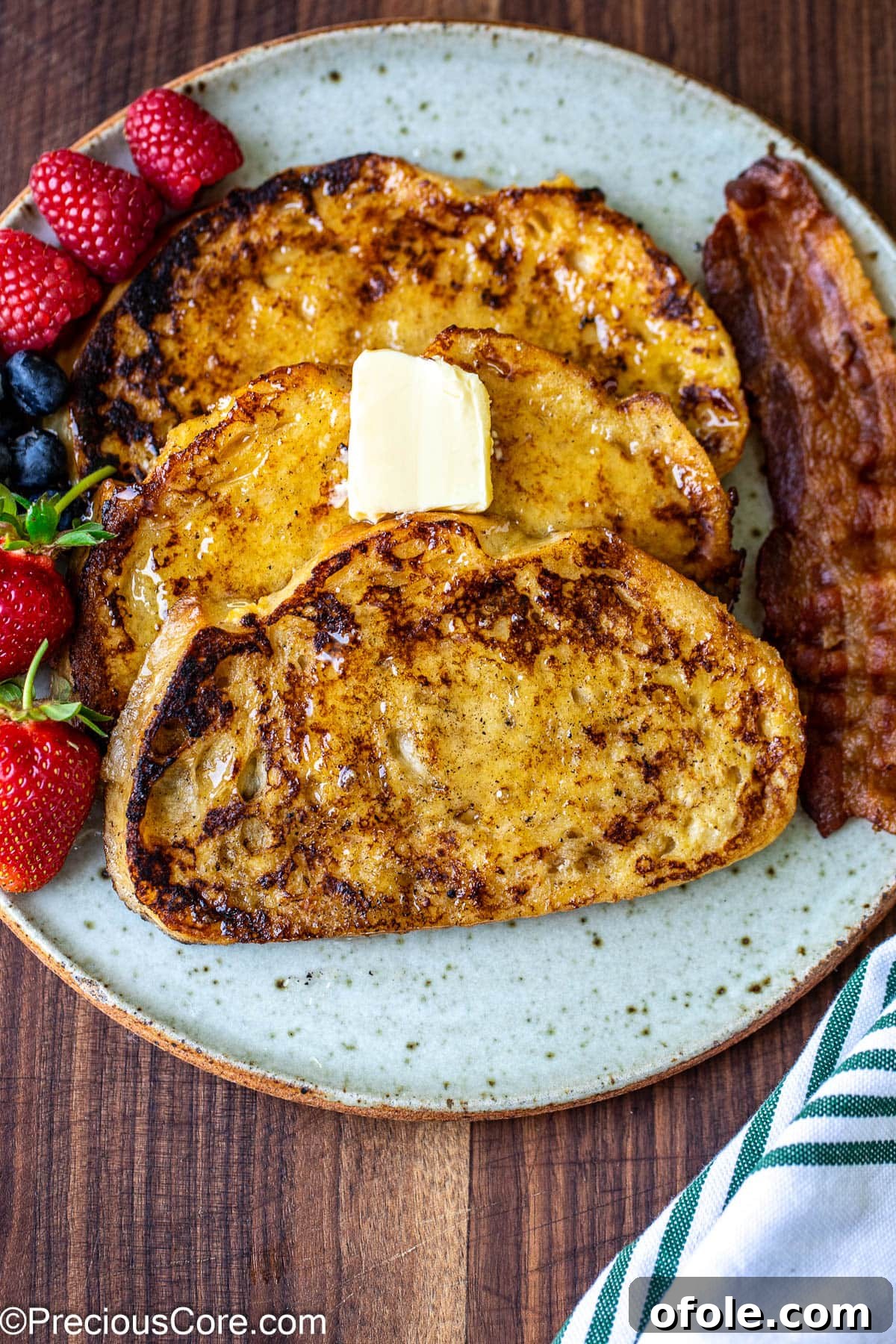 Elevated Sourdough French Toast 3 A serving of three Sourdough French toast slices, artfully arranged on a plate, complemented by fresh fruit and crispy bacon on the side.