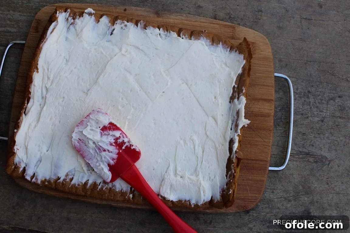 Spiced Pumpkin Swirl Cake 16 A red offset spatula expertly spreading a thick layer of creamy cream cheese filling across the flat pumpkin cake, ensuring even coverage before rolling.