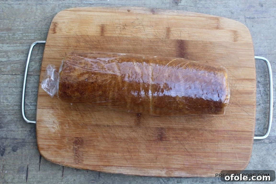 Spiced Pumpkin Swirl Cake 17 The pumpkin cake, now generously filled with cream cheese, is expertly rolled into a cohesive log, ready for chilling, on a wooden cutting board.