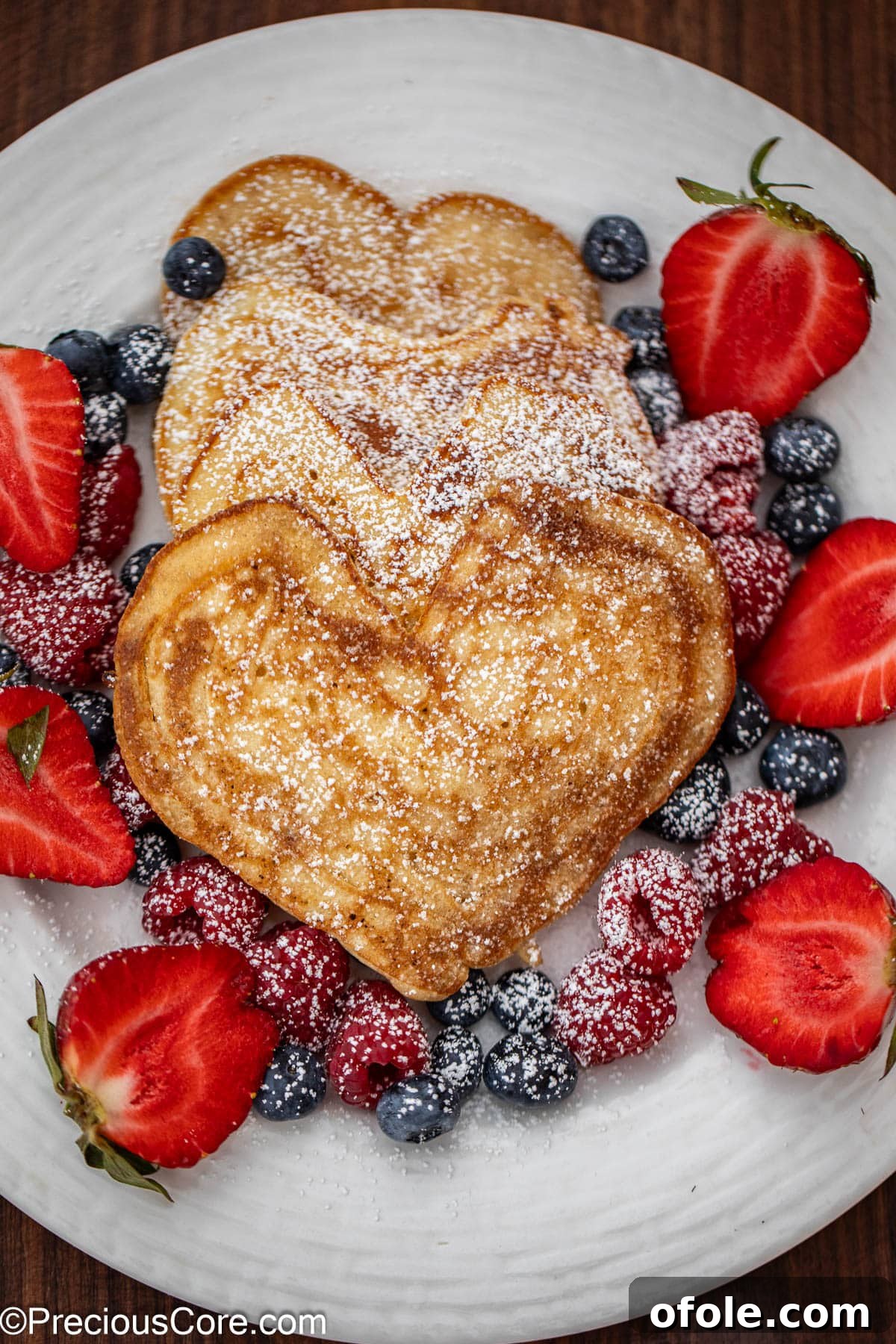 Sweetheart Flapjacks 2 A stack of fluffy heart shaped pancakes artfully arranged on a white plate, garnished with vibrant fresh berries and a drizzle of golden maple syrup.