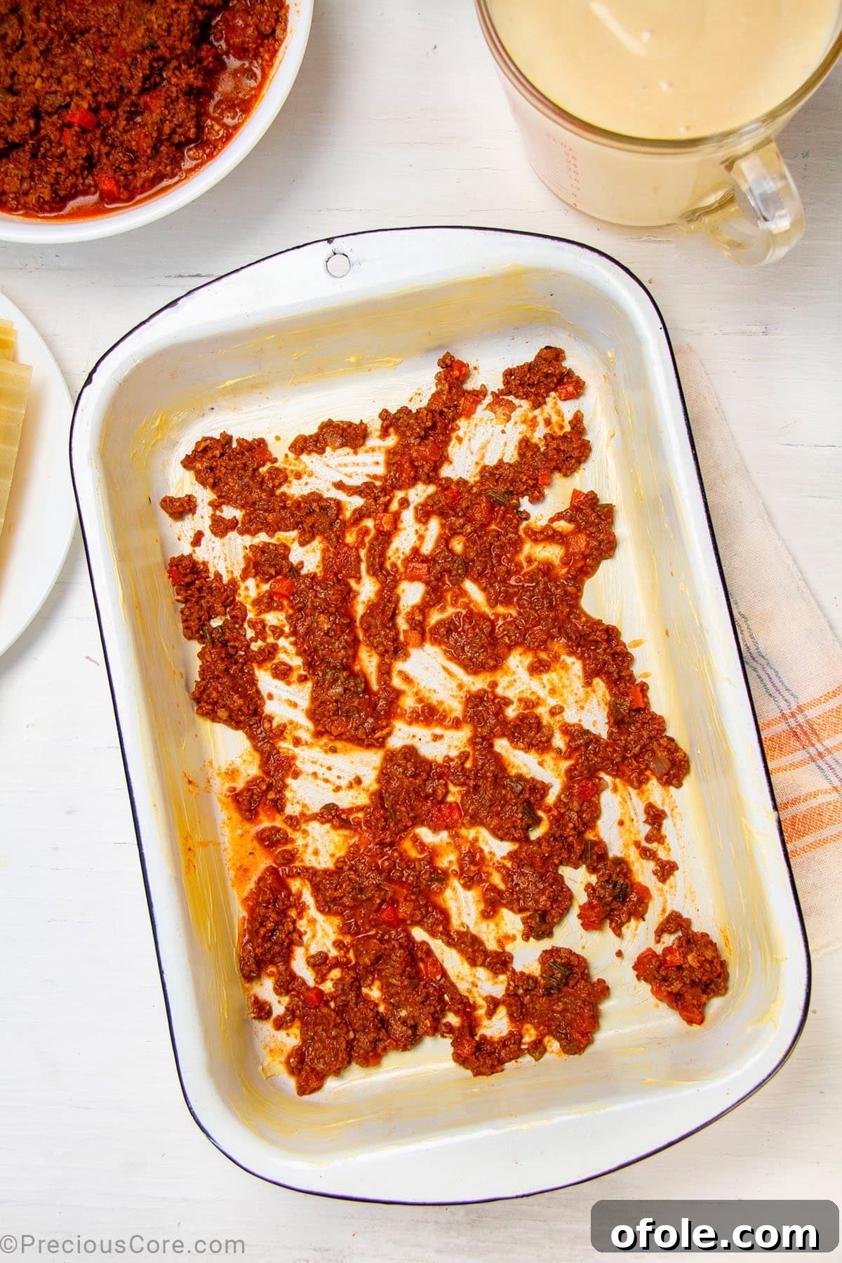 Creamy Béchamel Lasagna 12 A thin layer of meat sauce evenly spread across the bottom of a glass baking dish, preparing the base for lasagna layering.