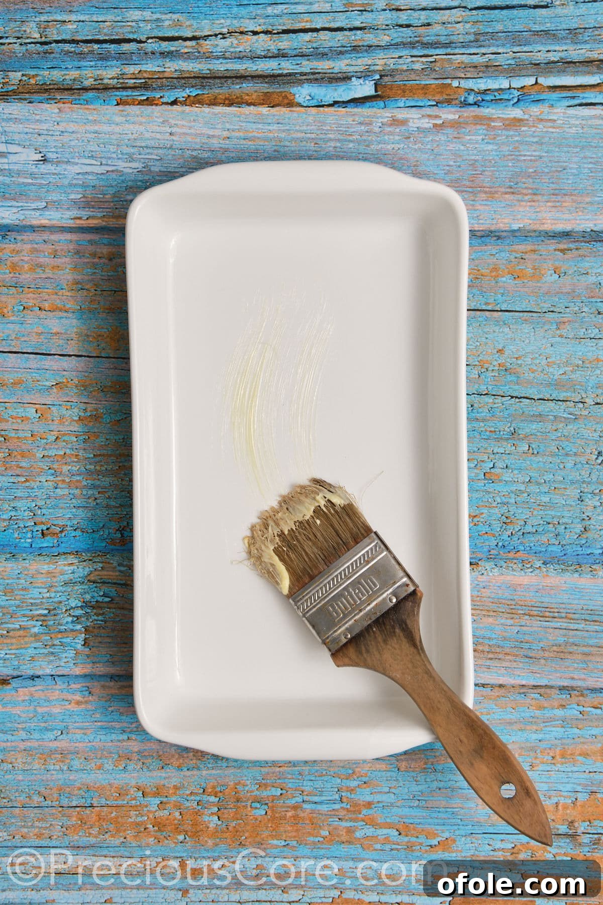 A hand brushing softened butter into a 9x13 inch casserole dish, ensuring all sides are coated for non-stick baking.