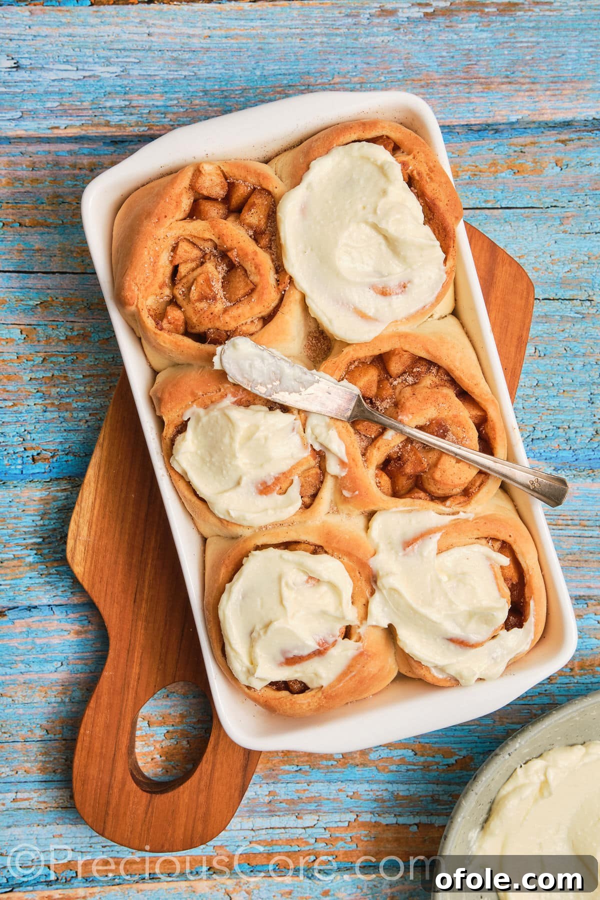 A spatula spreading thick, white cream cheese frosting over warm, golden-brown apple cinnamon rolls fresh out of the oven.
