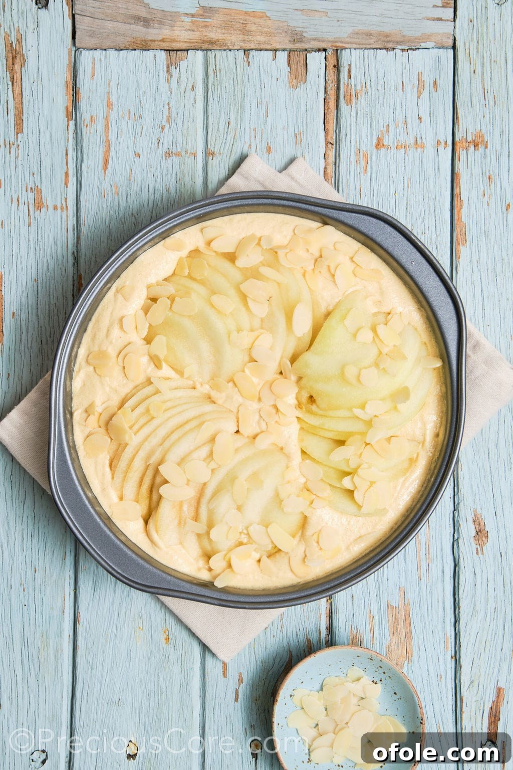 Cake batter in a pan, beautifully topped with fanned-out pear slices and a generous sprinkling of almond flakes, ready for baking.