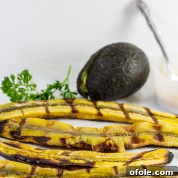 Grilled plantains with an avocado. A close-up shot of golden-brown plantain halves with distinct grill marks, served alongside creamy green avocado slices.
