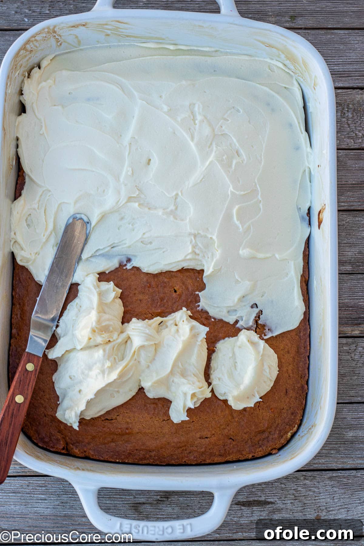 Spiced Pumpkin Cake with Luscious Cream Cheese Frosting 6 Hands spreading creamy white cream cheese frosting evenly over a baked pumpkin cake in a pan.