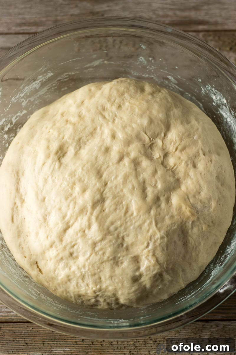 Glass bowl filled with soft, risen beignet dough, covered with plastic wrap.
