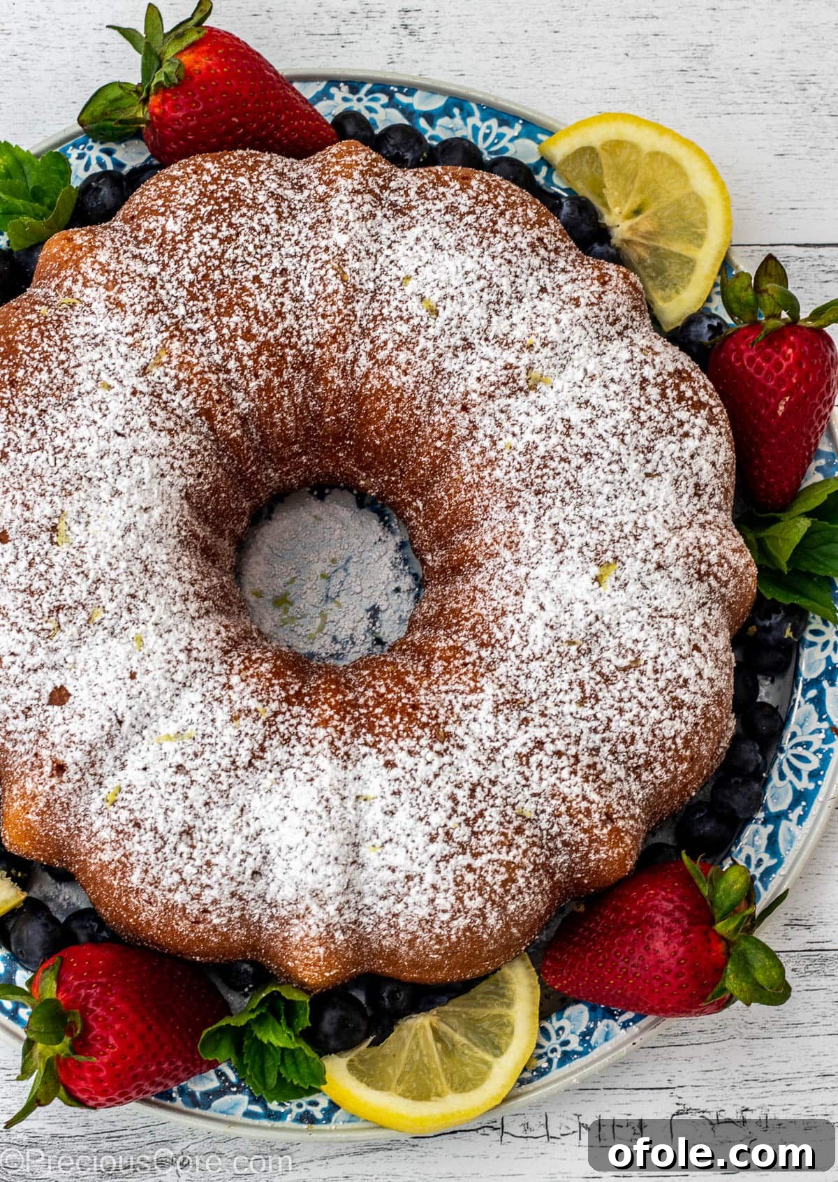 Zesty Southern Lemon Pound Cake 9 A freshly baked bundt cake lightly dusted with powdered sugar, surrounded by vibrant mixed berries on a white surface.