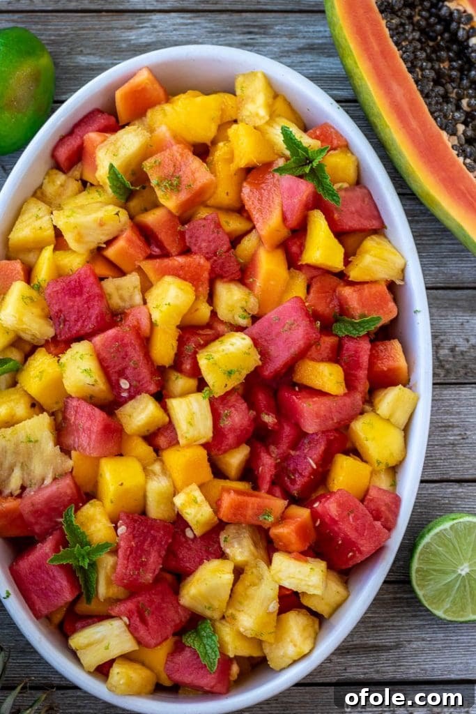 Fresh Ingredients for Tropical Fruit Salad Tropical fruit salad in a white ceramic bowl, with a halved papaya and whole limes neatly arranged beside it on a wooden surface.