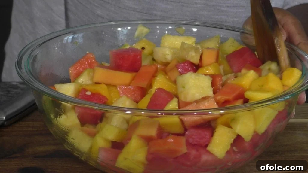 Mixing Tropical Fruit Salad with Dressing A hand gently mixing a large bowl of tropical fruit salad with a wooden spoon, ensuring the dressing coats all the vibrant fruit pieces.