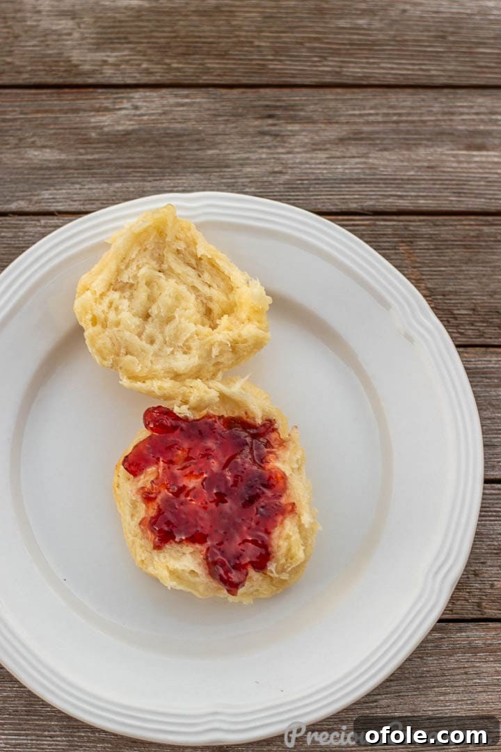A single warm buttermilk biscuit on a white plate, cut open with jam generously spread on top, ready to eat.