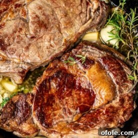 Two ribeye steaks in a cast iron skillet with rosemary, thyme, and garlic, perfectly seared.